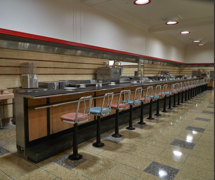 An empty vintage diner counter with colorful stools, coffee cups, and retro kitchen equipment, under bright overhead lights.