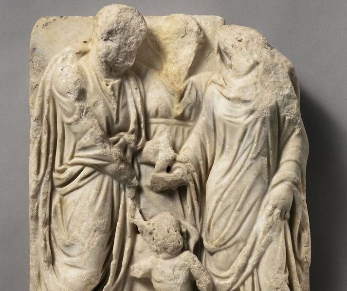 Weathered marble relief showing a family group with a child; the adult figures are mostly headless but wear flowing garments, suggesting a domestic or funerary scene.