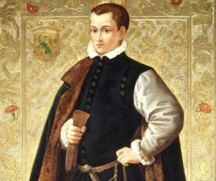 Lord Guildford Dudley stands in dark formal dress with a fur-trimmed cloak, poised before a patterned backdrop bearing a family crest.