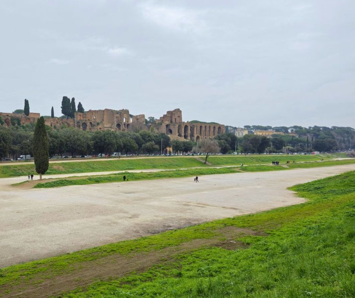 Wide, grassy expanse of the ancient Circus Maximus with scattered visitors, bordered by trees and Roman ruins rising on the Palatine Hill in the background.