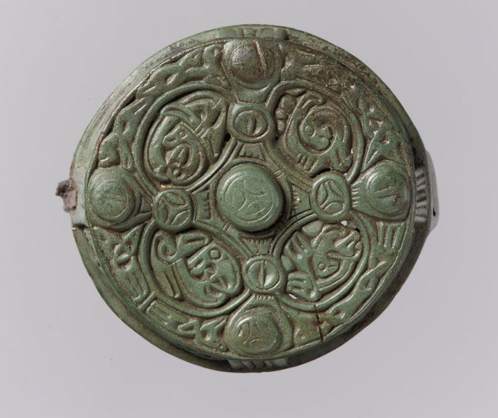 A round metal brooch with intricate carvings, possibly of medieval or Viking origin. It features symmetrical patterns, circular motifs, and stylized animal or script designs, with a weathered green patina.