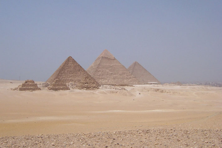 The Pyramids of Giza standing prominently in the Egyptian desert, with their iconic triangular shapes rising against a clear blue sky and surrounded by vast sandy terrain.