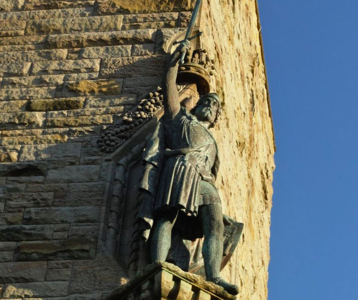 A bronze statue of William Wallace, standing tall with a raised sword. The statue is mounted on the Wallace Monument, against a clear blue sky.