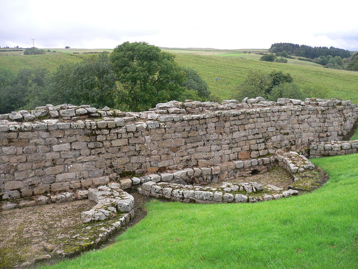 Section of an old stone wall with semicircular foundations set in green grass, surrounded by rolling countryside.