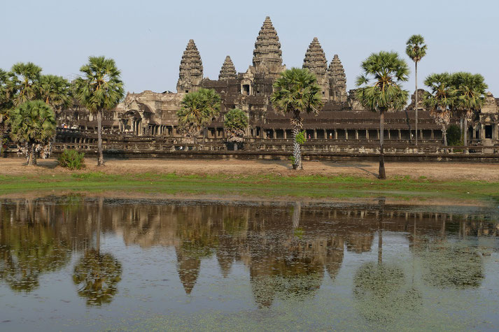 Angkor Wat, the iconic Khmer temple complex in Cambodia. Its majestic towers rise above a reflective pool, surrounded by palm trees and bathed in soft daylight.