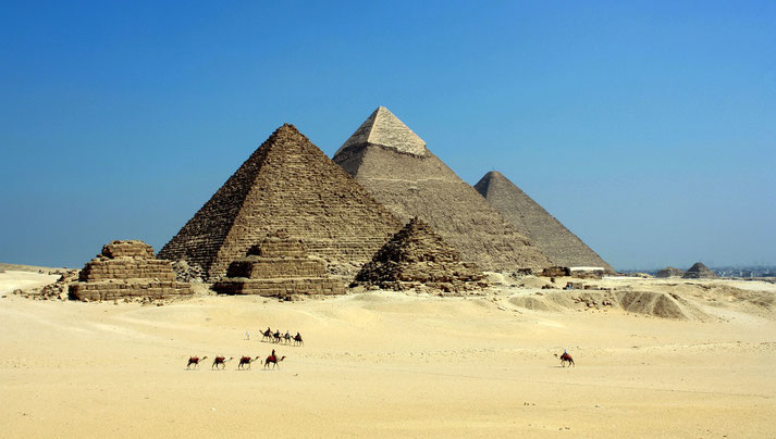 The Pyramids of Giza under a clear blue sky, with camel riders traversing the desert. Smaller pyramids and ruins surround the iconic structures, highlighting their ancient grandeur.