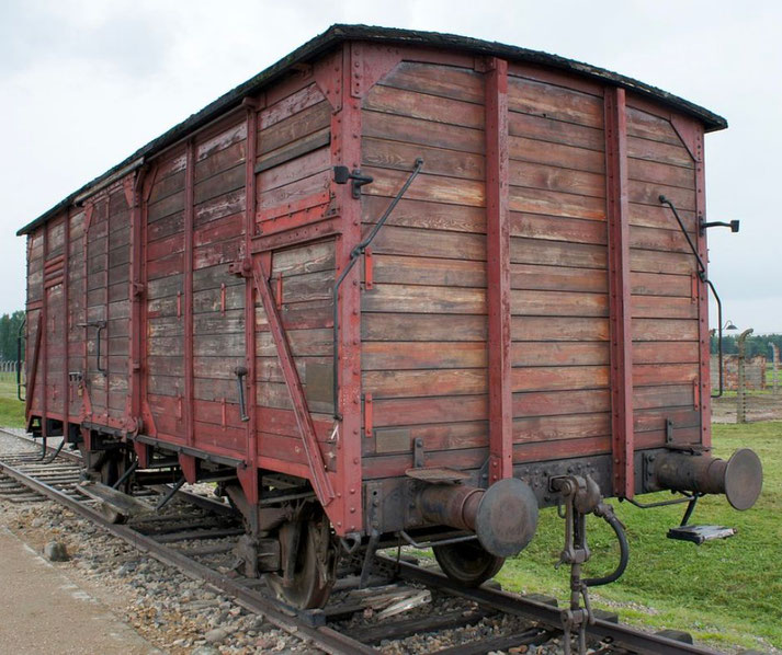 A weathered wooden boxcar sits on railway tracks near a historic site, once used for transporting people during wartime.