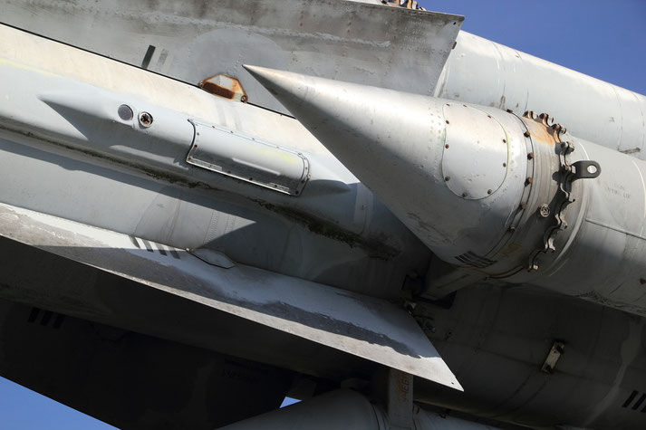 Close-up of a metallic missile nose cone with fins and rusted bolts against a clear blue sky.