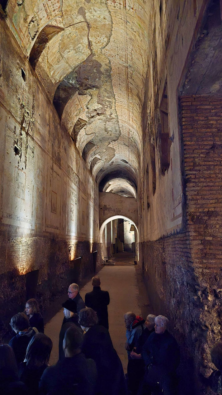 Visitors explore a long, dimly lit ancient corridor with a vaulted ceiling, faded frescoes, and arched doorways, showing signs of age and partial restoration.