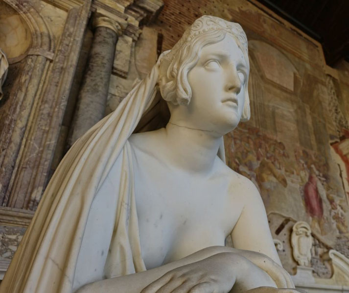 Marble statue of a veiled woman with a solemn expression, partially draped, set against a background of classical frescoes and columns.