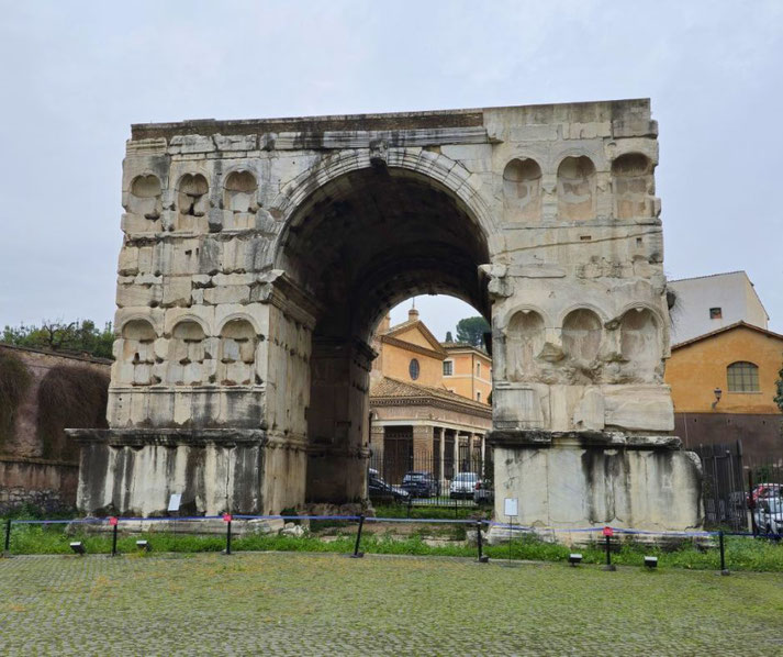 Ancient Roman triumphal arch with a central vaulted passage, weathered stone, and decorative niches, standing on cobbled ground with buildings visible through the arch.
