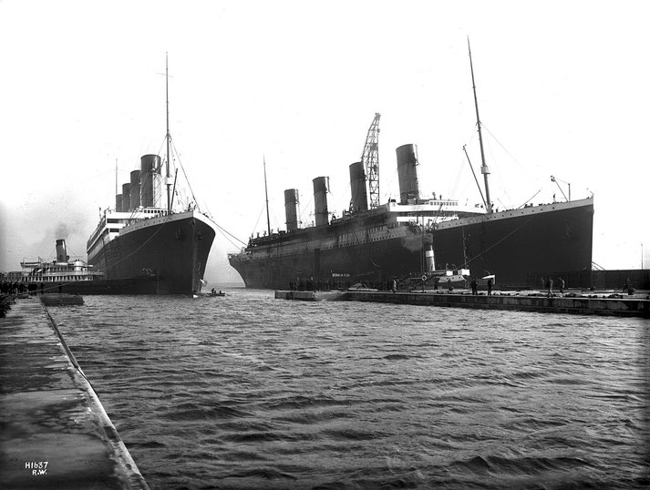 Two massive ocean liners docked side by side, featuring multiple smokestacks and a bustling pier with workers and small boats nearby, highlighting the grandeur of early 20th-century shipbuilding.