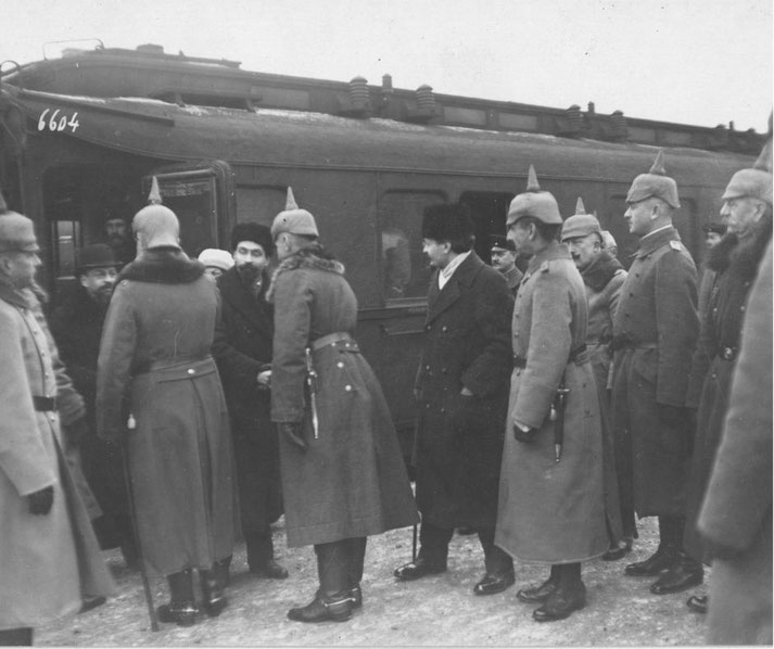 Black-and-white photo of uniformed soldiers in spiked helmets greeting civilians beside a train carriage at a station, likely during an early 20th-century military or diplomatic arrival.