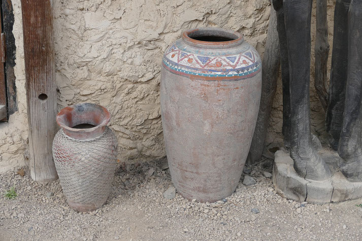 Two large weathered clay pots, one with geometric painted patterns and the other covered in mesh, rest on gravel against a textured wall.