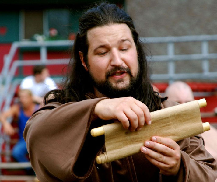 A bearded historical reenactor in a brown robe reading from a scroll. He gestures expressively, possibly portraying an ancient poet, philosopher, or lawgiver in an outdoor setting.