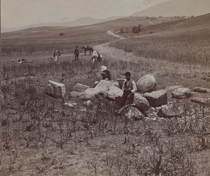 Sepia-toned photograph of 19th-century archaeologists at a rural excavation site, surrounded by scattered ancient stone ruins and open landscape.