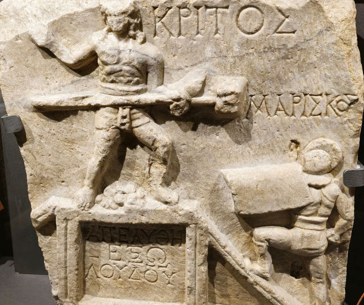 Stone relief carving showing a net-armed retarius raising a trident against a helmeted secutor on a stepped platform with Greek inscriptions.