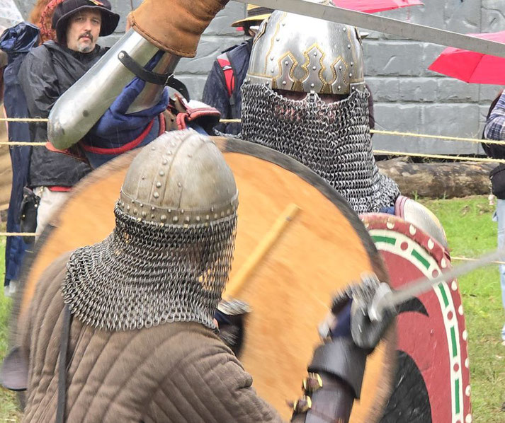 A live-action medieval reenactment showing two armored fighters in chainmail and helmets clashing with swords and shields, surrounded by onlookers and props that evoke a historical setting.