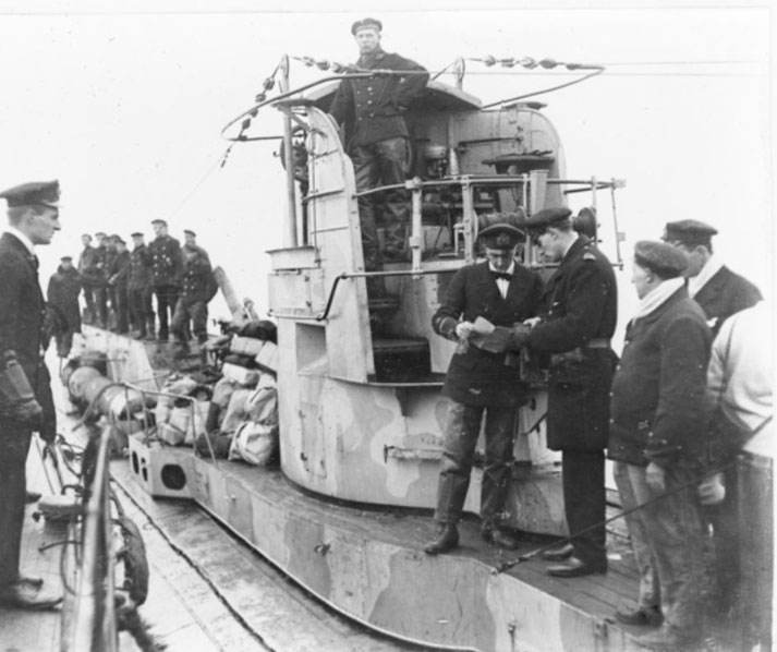 Sailors and officers gather on the deck and conning tower of a submarine, some reviewing documents while others stand in formation.