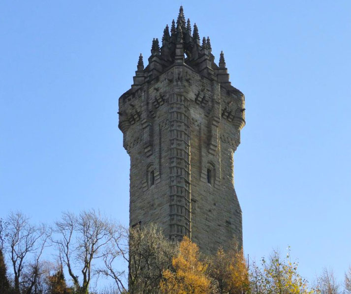The top of the Wallace Monument, a tall, gothic-style stone tower with pointed spires, set against a clear blue sky and surrounded by autumn trees.