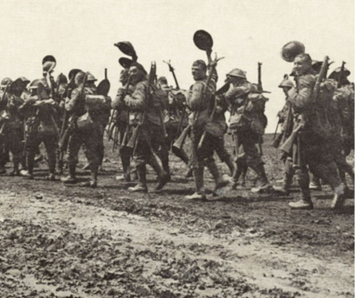 A column of World War I soldiers marches on a muddy road, carrying rifles, packs, and gear including shovels and helmets raised.