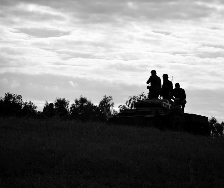 Silhouetted soldiers stand on and around a tank atop a hill, with trees and a cloudy sky forming the backdrop.