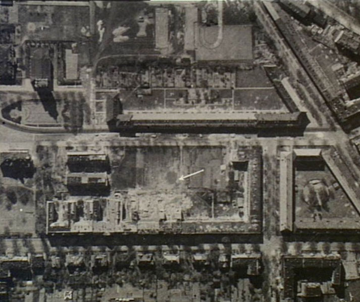 Aerial photo of bombed urban area with buildings in ruins; arrow points to a specific site among the destruction.