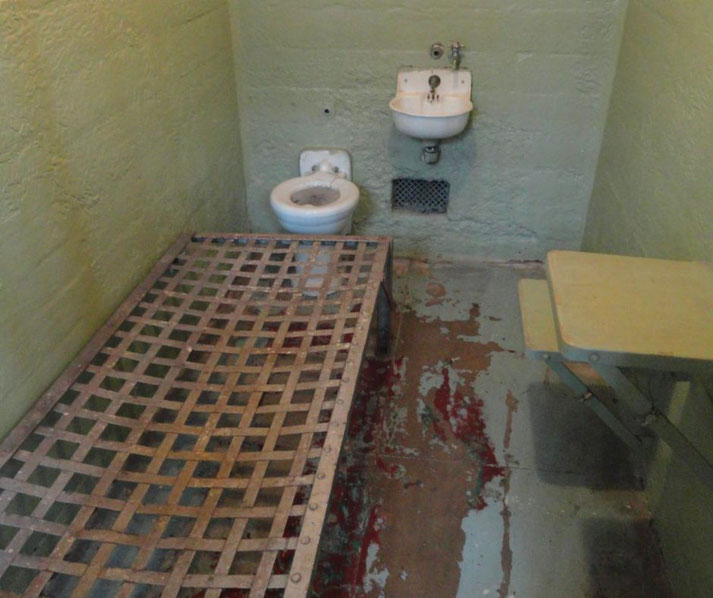 A small prison cell with a metal bed frame, toilet, sink, and wall-mounted table on a worn, paint-chipped floor.