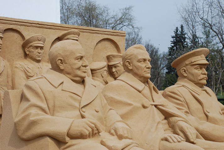 Sand sculpture of seated military and political leaders in uniform, featuring detailed facial expressions and attire, displayed outdoors near bare trees.