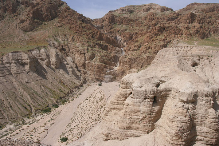 A rugged desert canyon with steep, layered rock formations and dry terrain, dotted with sparse vegetation under a clear blue sky, evoking an arid, remote atmosphere.