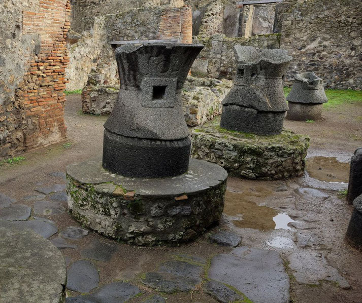 Stone mills in the Bakery of Modestus at Pompeii, used for grinding grain. The setting includes brick and stone walls, mossy bases, and puddles on the ground, reflecting ancient craftsmanship.