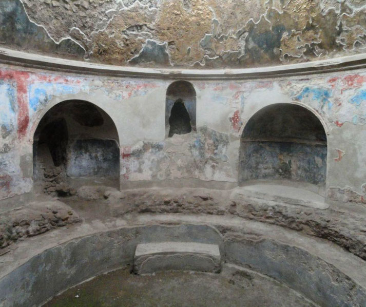 The interior of the baths at Pompeii, featuring semi-circular niches, faded frescoes with red and blue accents, and a worn stone bench, showcasing ancient Roman bathing architecture.