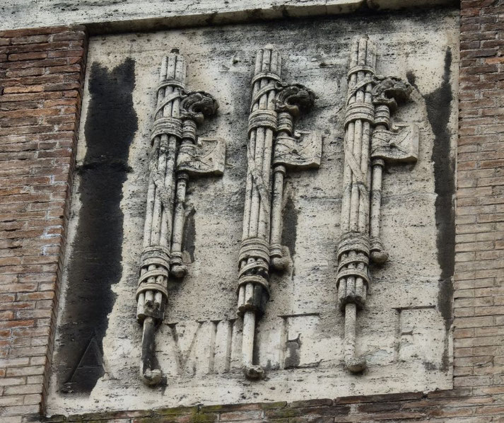 Stone relief of three Roman fasces—bundles of rods with axes—carved into a wall, symbolizing authority and power, with faint Latin inscriptions below.