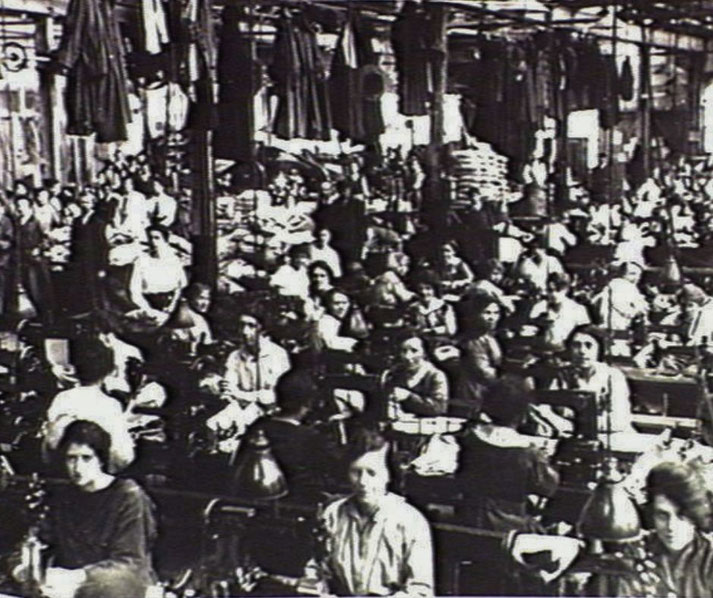 Crowded factory filled with women working at sewing machines, surrounded by hanging garments and dim lighting, likely from the early 20th century.