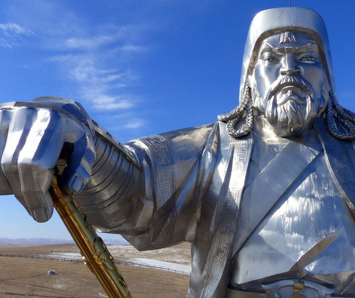 A massive metallic statue of a warrior with braided hair and a stern expression holds a golden staff, set against an open landscape and blue sky.