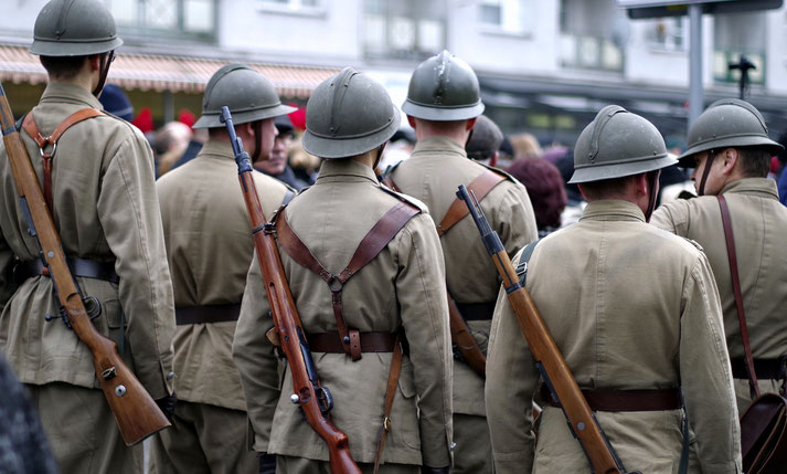 Group of soldiers in vintage World War I-style uniforms and helmets, carrying rifles on their backs, seen from behind during a public event or reenactment.