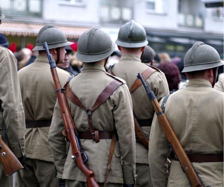 Group of soldiers in vintage World War I-style uniforms and helmets, carrying rifles on their backs, seen from behind during a public event or reenactment.