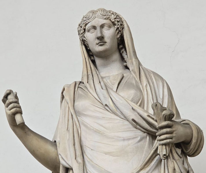 Marble statue of a robed woman with a veil over her head, holding scrolls in one hand and a cylindrical object in the other.