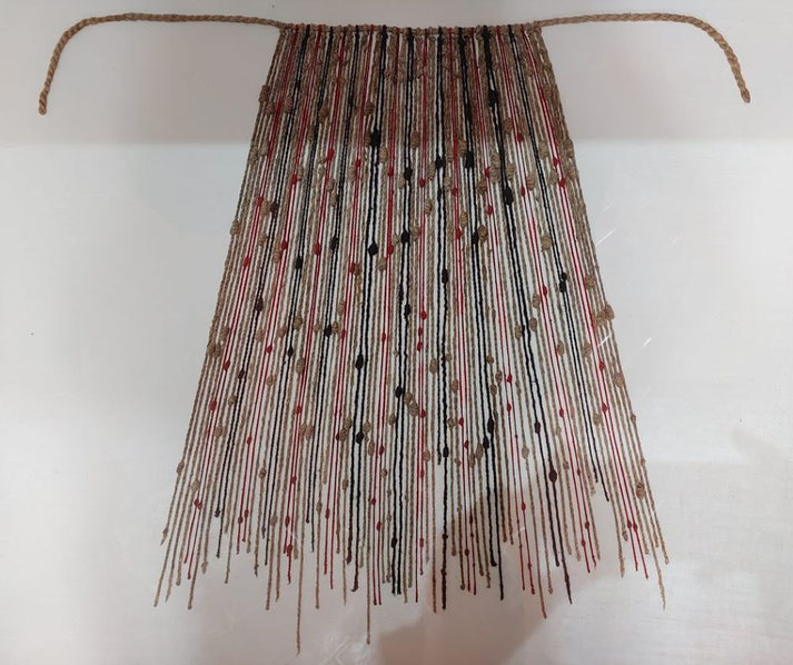 A hanging textile piece made of multicolored yarn strands in beige, red, and black, suspended from a twisted rope. The strands have knots and varying lengths, creating a textured, tapering pattern.