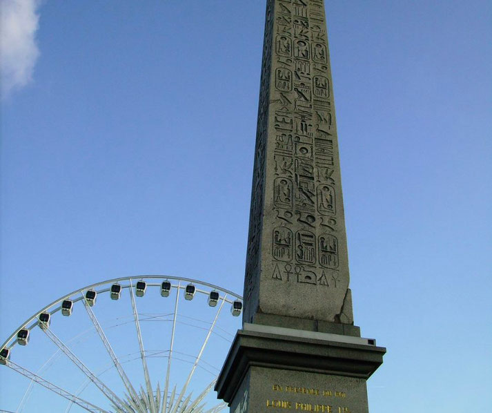 An ancient Egyptian obelisk with hieroglyphs stands tall in front of a modern Ferris wheel under a blue sky.