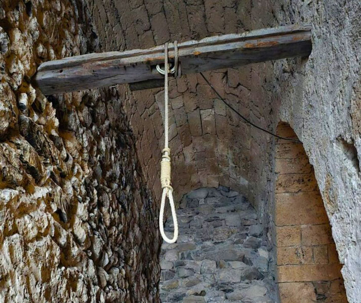 A weathered noose hangs from a wooden beam in a medieval stone passageway. The rough walls and dim atmosphere evoke a grim historical setting, likely linked to justice or punishment in the past.