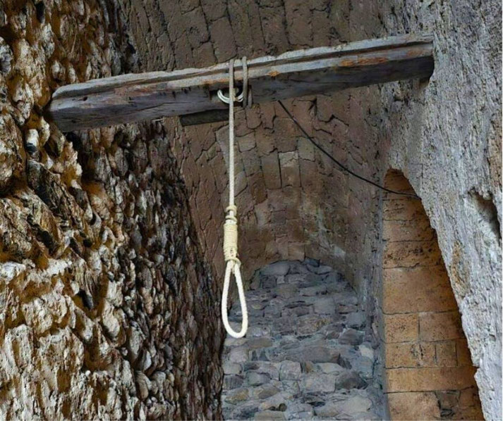 A weathered noose hangs from a wooden beam in a medieval stone passageway. The rough walls and dim atmosphere evoke a grim historical setting, likely linked to justice or punishment in the past.