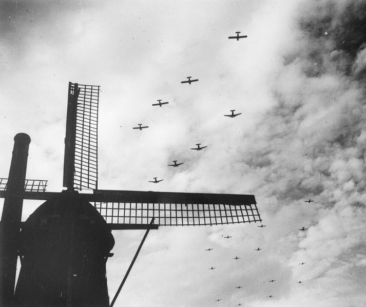 Silhouetted windmill in foreground with a large formation of military aircraft flying overhead in cloudy skies.