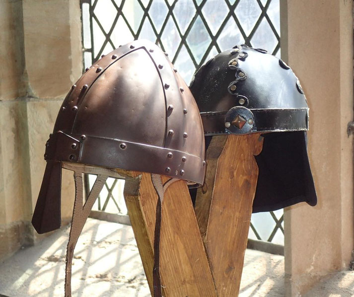 Two recreated Viking helmets, one made of bronze and the other of blackened steel, featuring riveted designs and simple, functional shapes, displayed on wooden stands in a medieval-style setting.
