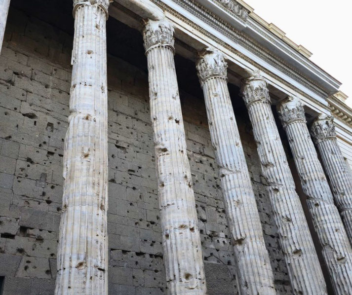 The weathered Corinthian columns of the Temple of Hadrian in Rome. The stone facade bears visible damage.