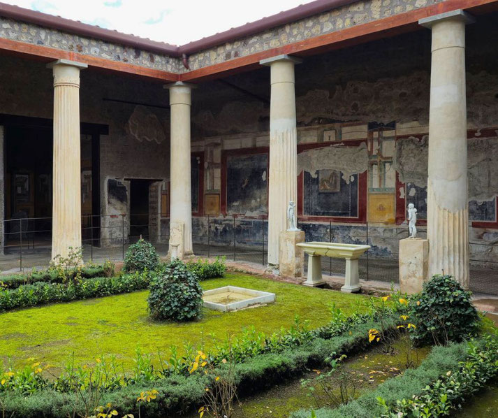The garden of the House of the Vettii in Pompeii, featuring a lush courtyard with hedges, statues, a small fountain, and frescoed walls framed by elegant columns under a covered walkway.