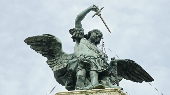 Close-up of a bronze angel statue with raised sword and spread wings atop a stone pedestal under a cloudy sky.