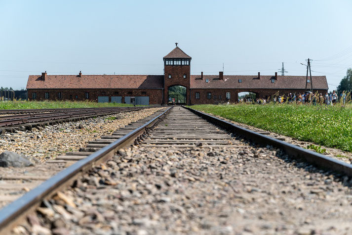 The entrance to Auschwitz-Birkenau concentration camp, showing the infamous railway tracks leading to the brick gatehouse under a clear sky, symbolizing a solemn reminder of history.