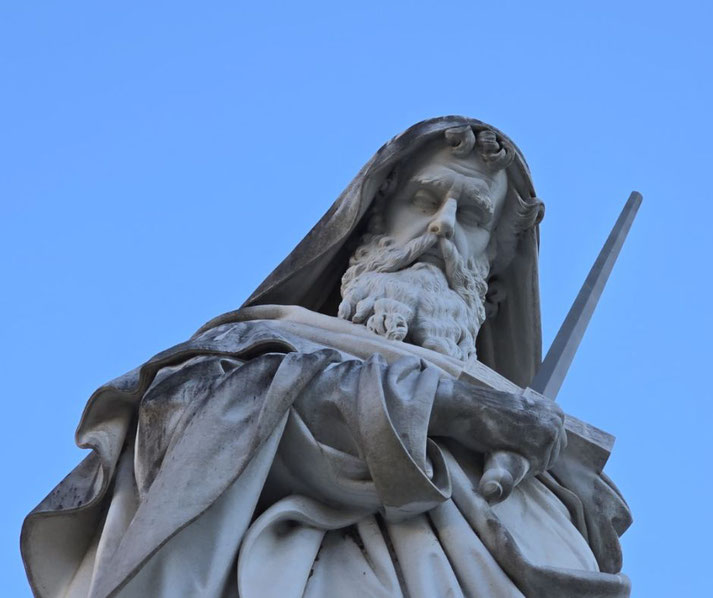 Marble statue of a bearded man in a hooded robe holding a sword, viewed from below against a clear blue sky.