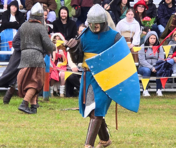 A medieval reenactment with two armored fighters wielding swords and shields.